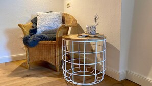 Une pièce d’angle avec un fauteuil en osier, une petite table d’appoint ronde en fil de fer blanc et une petite figurine blanche.