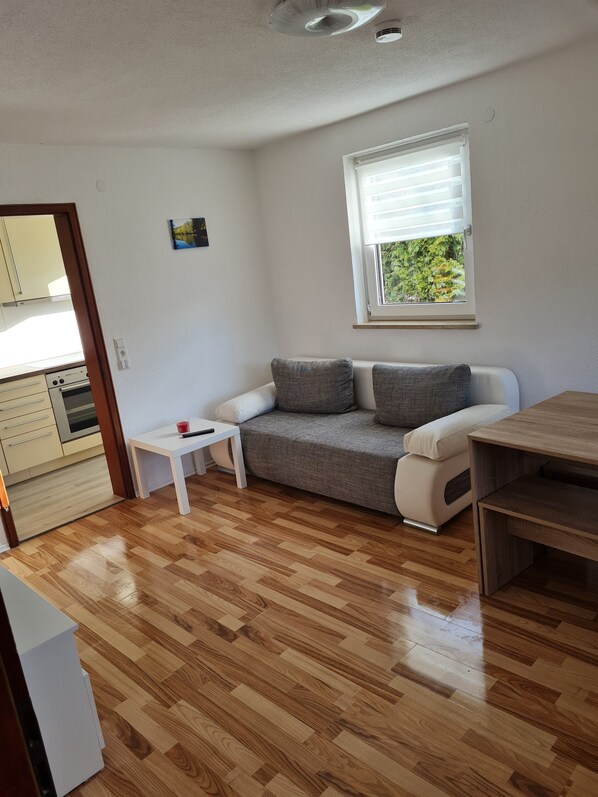 A compact living space with a kitchenette, a sofa with cushions, a small table, and a window with blinds.
