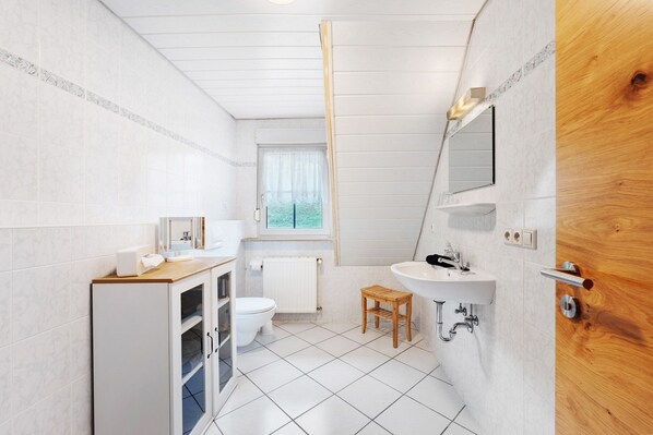 Un baño con piso de baldosas blancas, un inodoro, un lavamanos, un taburete de madera y una ventana con vistas a la vegetación.