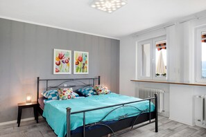 A bedroom with a bed, two wall-mounted paintings, a bedside table with a lamp, a window with curtains, and a radiator.
