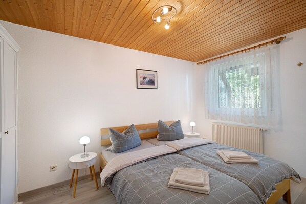 A bedroom with a wooden ceiling, a bed with grey bedding, a bedside table with a lamp, and a window with sheer curtains.