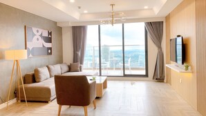 A modern living room with a sofa, armchair, and coffee table, large windows with a city view, and a mounted TV. - Ha Tay Millennium Apartments - Free Gym Pool (Hanoi)