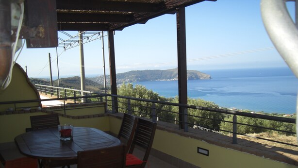 A covered patio with a table and chairs, overlooking a coastal landscape with hills and the sea.