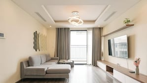 Un salon moderne avec un canapé d’angle, une table basse ronde, un téléviseur à écran plat fixé sur une console en bois et une grande fenêtre avec des rideaux.