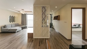 Un salon moderne avec un canapé d’angle gris, un plancher en bois, un élément de séparation décoratif blanc et une vue sur une chambre à coucher avec un lit et un téléviseur.
