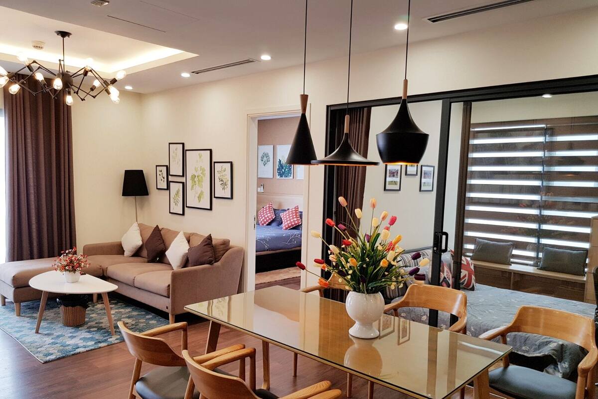 Un salon moderne avec une table à manger en verre, des chaises en bois, un canapé et une table basse. On peut y voir des tableaux encadrés au mur, un vase avec des fleurs et une grande fenêtre avec des stores.