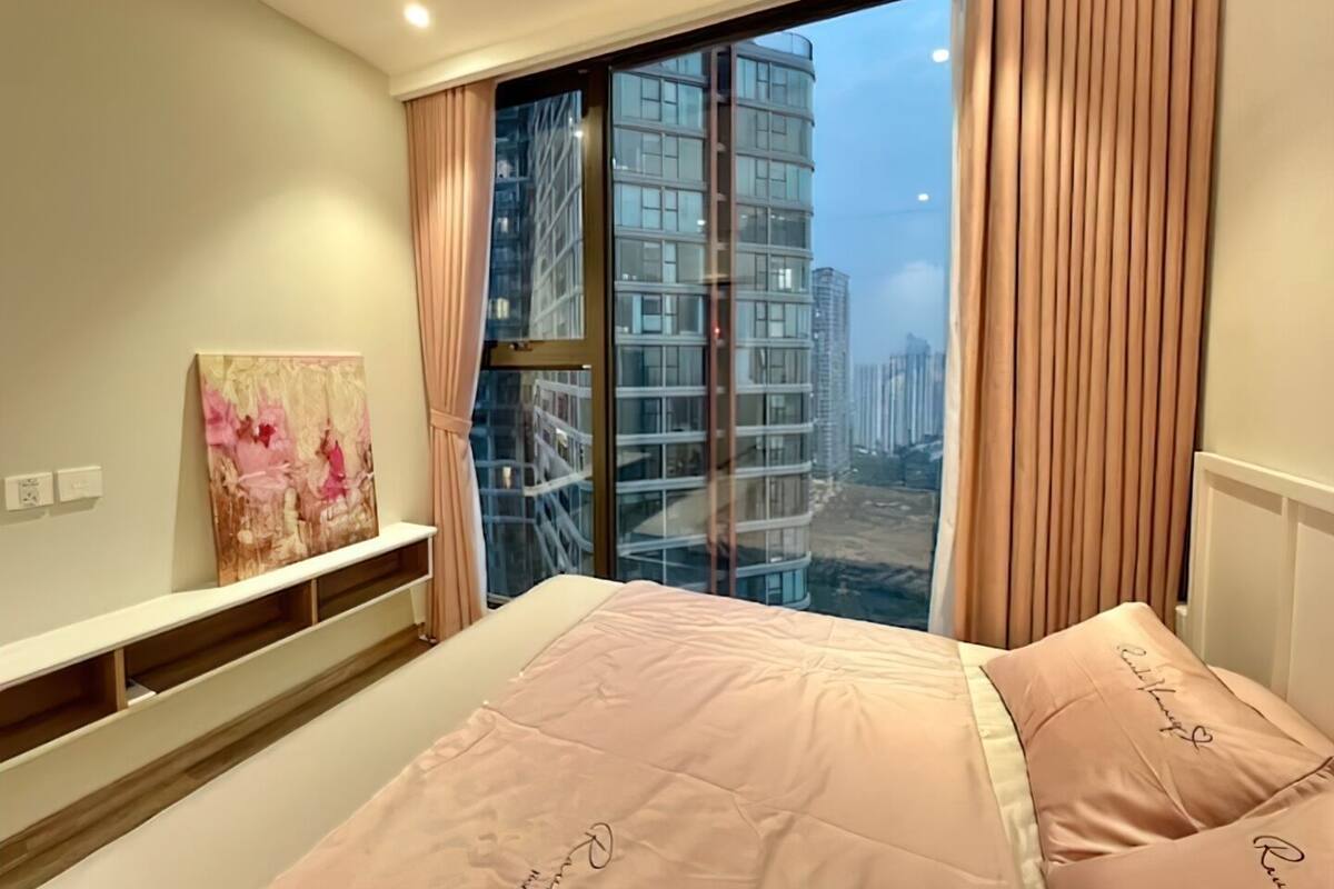 A bedroom with a large window, a bed with pink bedding, and a painting on the wall.