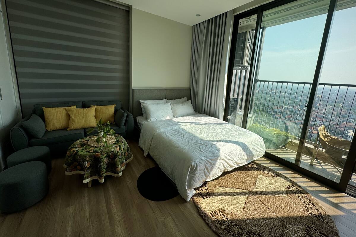 A modern bedroom with a large bed, a sofa, a small table, and a view of a cityscape through large windows.