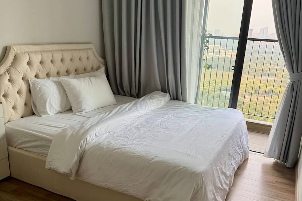 A bedroom with a large bed, grey curtains, and a view of greenery outside.