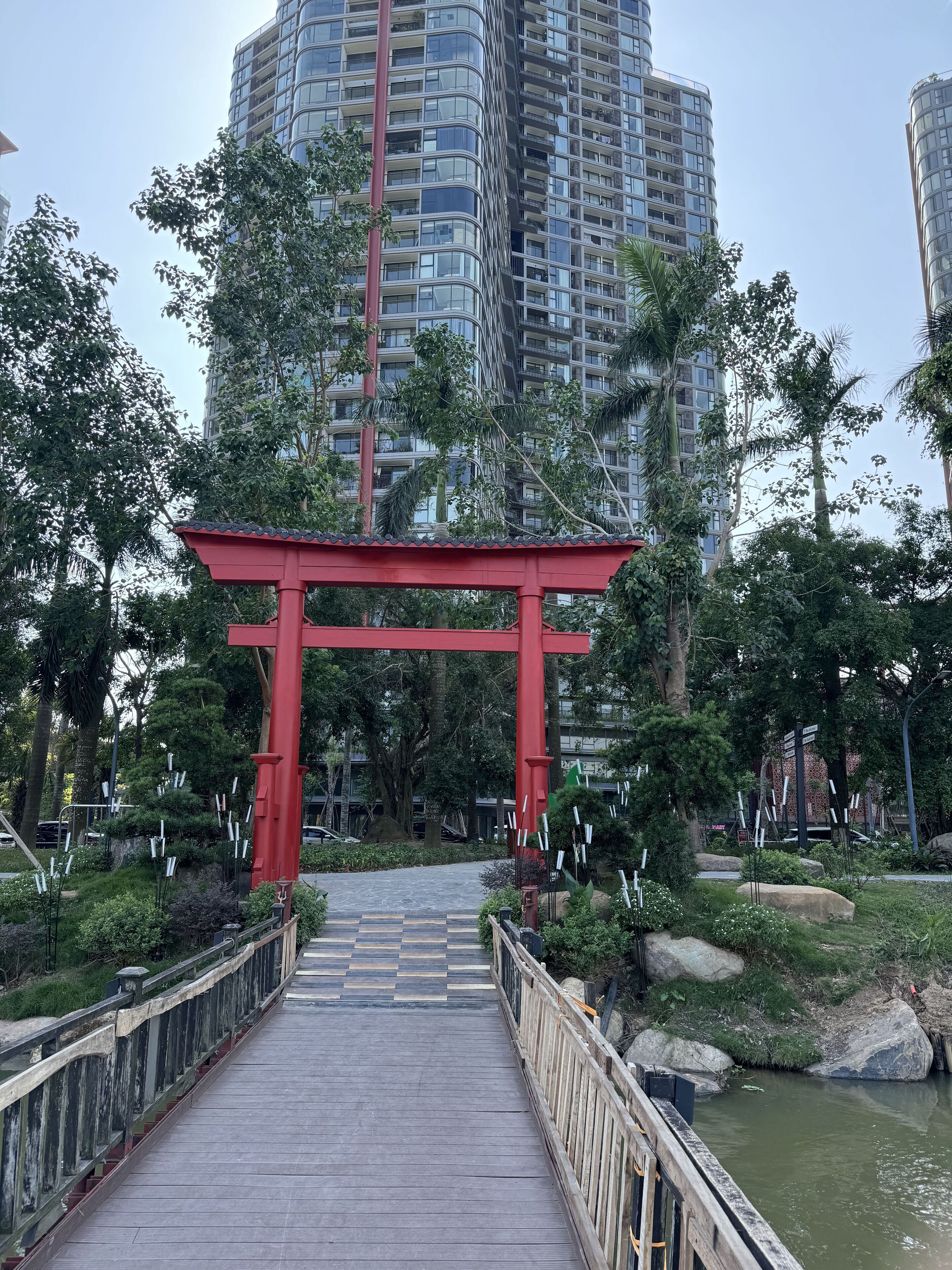 A pathway leading to a red torii gate, surrounded by greenery and a body of water, with tall buildings in the background.