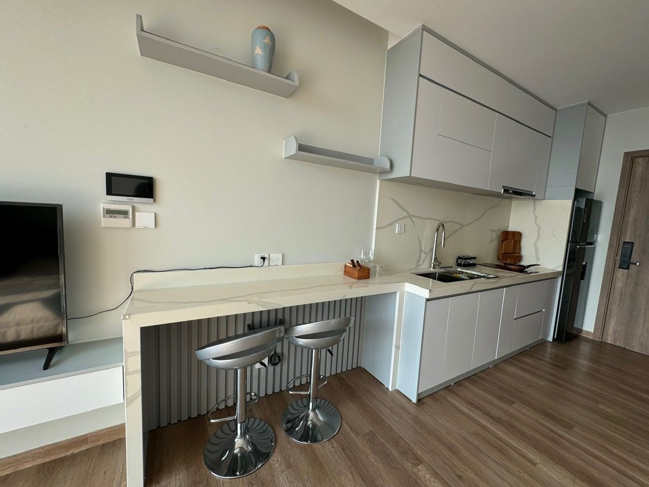 A modern kitchen with a white countertop, two bar stools, a built-in oven, and a TV mounted on the wall.
