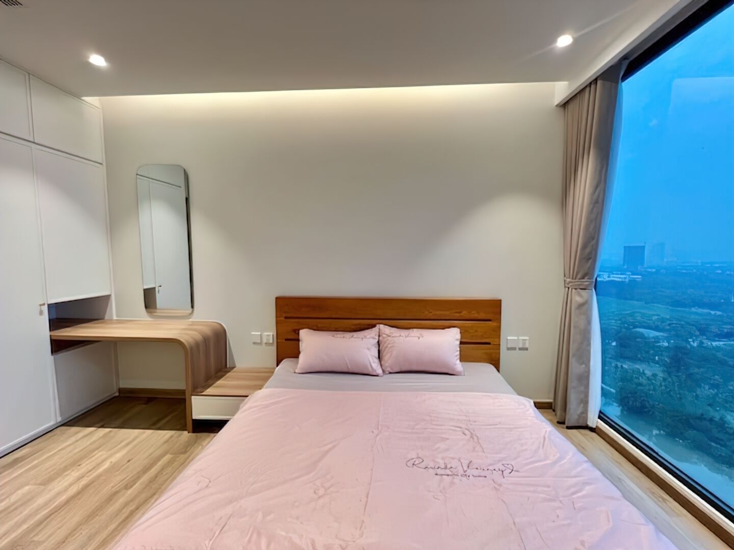 Un dormitorio moderno con una cama grande, cabecera de madera, un banco integrado, un ventanal con vistas a la ciudad y un espejo en la pared.