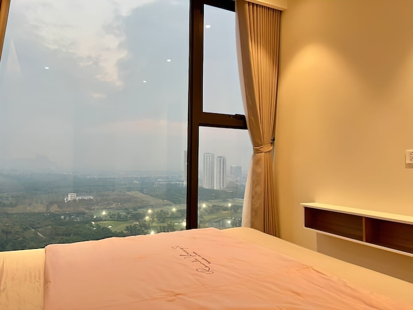 Ein Schlafzimmer mit großem Fenster, einem Bett mit rosa Bettdecke und Blick auf eine Stadtlandschaft.