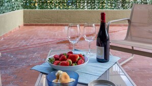 Une table en verre sur laquelle se trouvent une bouteille de vin, deux verres à vin, un bol de fraises, un panier de pain et une petite assiette. La scène se déroule sur un balcon, avec une haie verte et un bâtiment en arrière-plan.