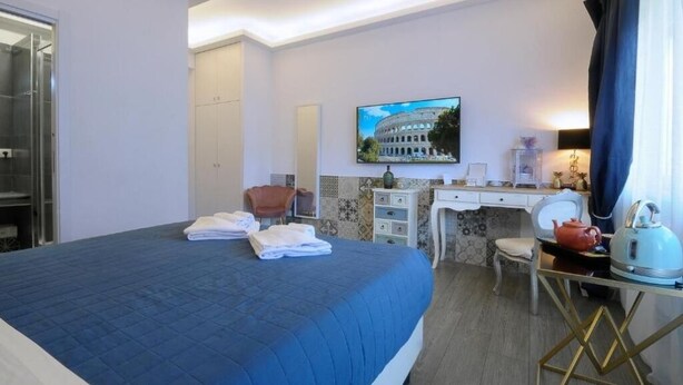 A hotel room with a large bed, a desk with a computer, a chair, and a television displaying a building.