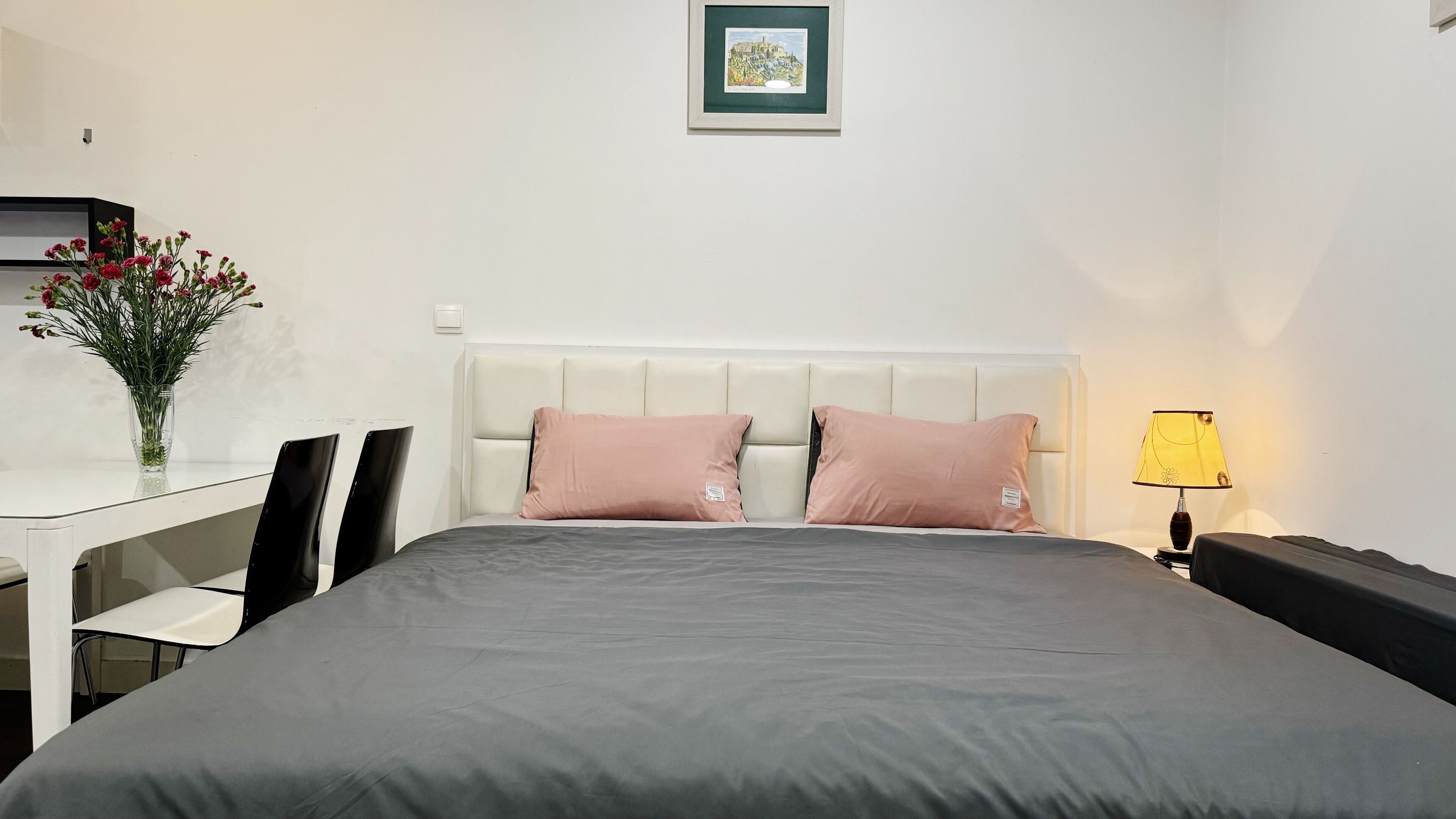 A neatly made bed with a grey bedspread and two pink pillows, a bedside table with a lamp, a chair, and a vase with flowers on a small table.