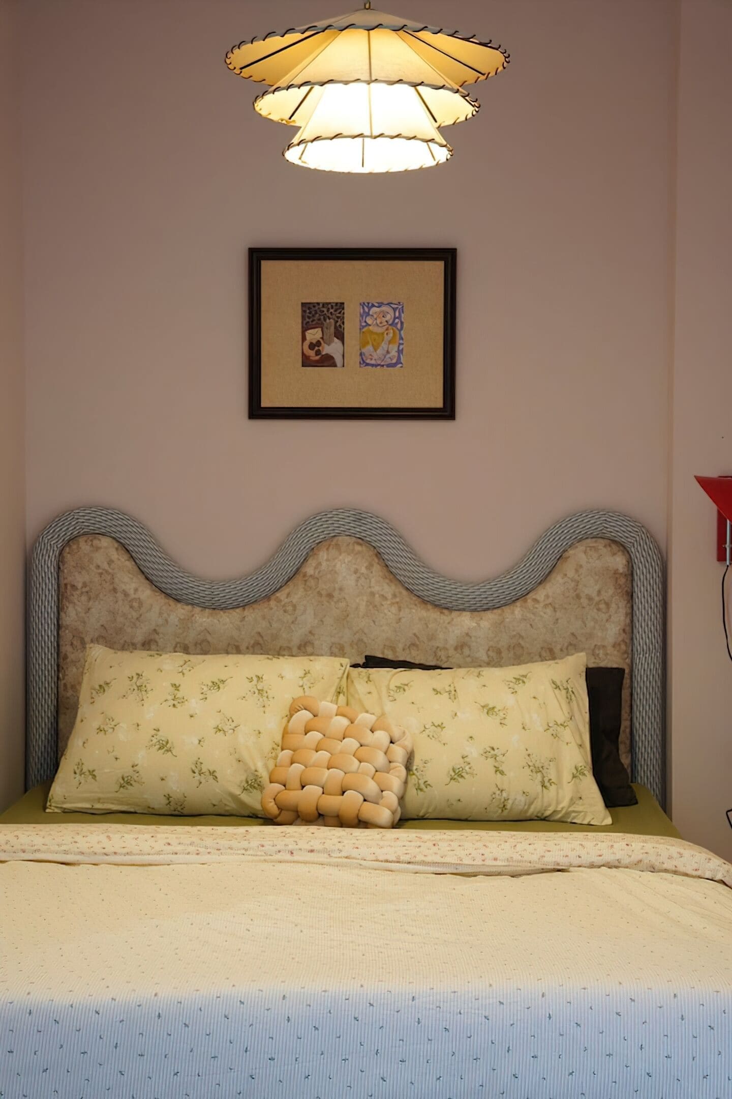 Un dormitorio con un cabecero ondulado, almohadas estampadas con motivos florales, una colcha de colores claros y un cuadro en la pared.