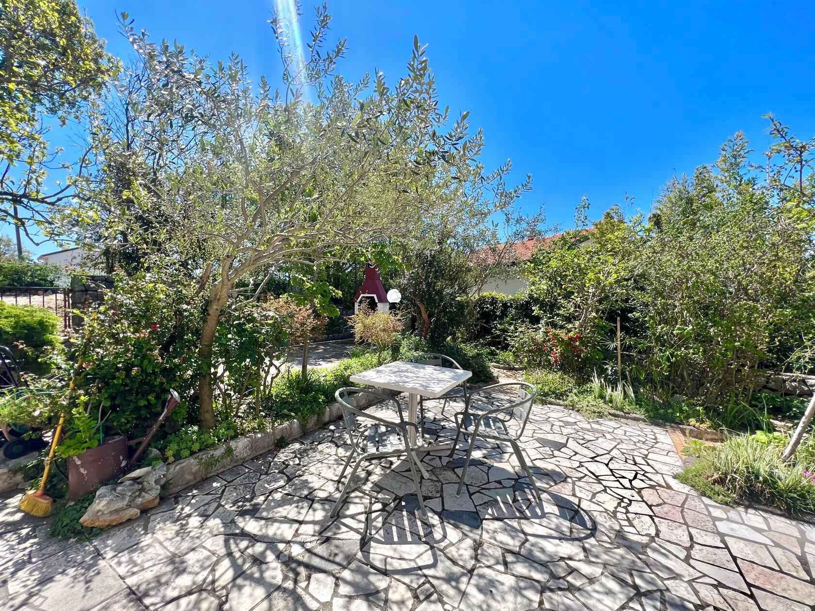 Un patio exterior con una mesa y sillas, rodeado de follaje y un sendero de piedra.