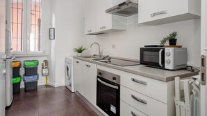 A modern kitchen with white cabinets, a built-in oven, a microwave, and a sink. There are three trash bins and a washing machine.
