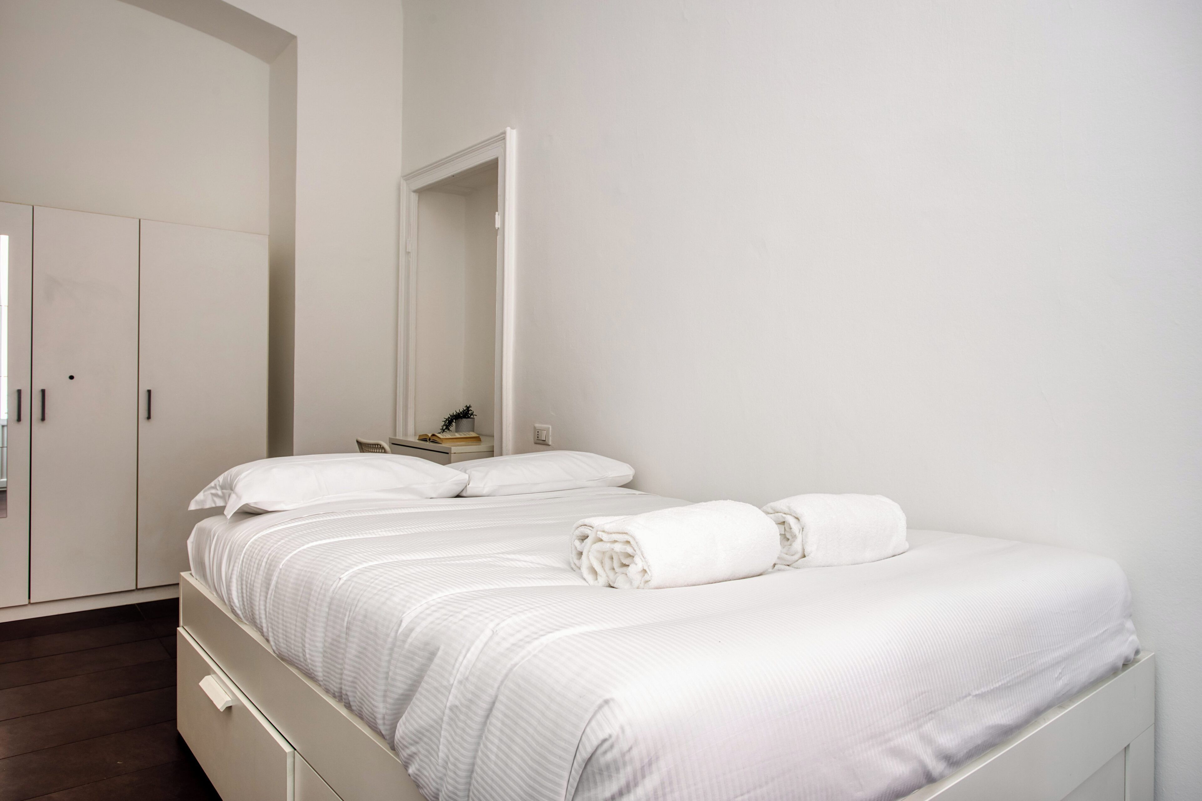 Una cama bien hecha con ropa blanca, un armario blanco y una mesita de noche con una planta.