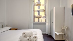 A hotel room with a bed, a chair, a wardrobe, and a window with a view of buildings.
