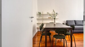 A dining area with a wooden table and chairs, a grey sofa, and a white wall with shelves.