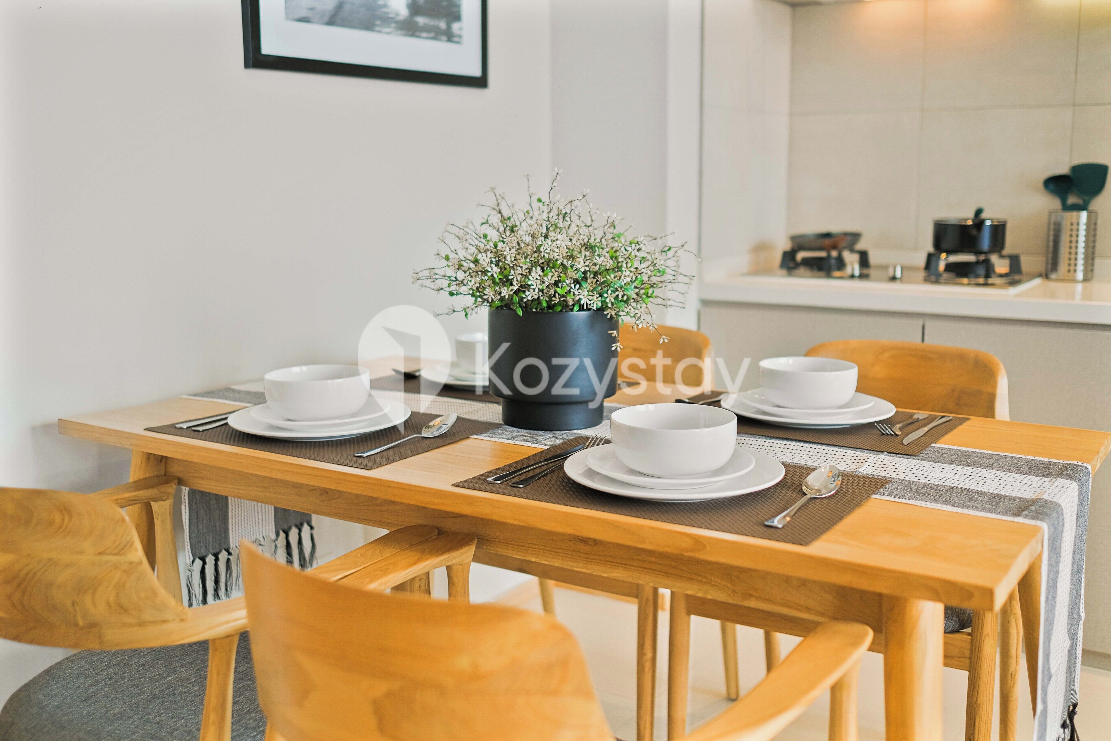 Une table à manger dressée avec de l’assiette, des bols et des couverts blancs, une plante en pot et un tableau encadré accroché au mur.