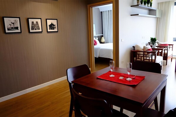 A hotel room with a wooden dining table, red placemat, and two wine glasses. There are three framed pictures on the wall and a bed with a white duvet in the background. - Mipec Riverside Long Bien Apartments (Hanoi)