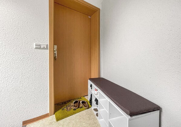 Un couloir avec une porte en bois, un porte-chaussures blanc avec des étagères marron, et un paillasson orné d’illustrations de chaussures.