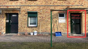 A brick building with a green entrance door labeled "Eingang/entrance" and a window with a metal grill. - Cozy & nature-loving apartment “Lichtblick” (Tschernitz)