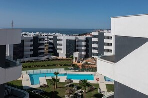 A modern building with a pool, surrounded by other buildings and a clear view of the sea.