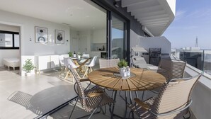 A modern interior with a round wooden table surrounded by wicker chairs, a glass dining table, and a kitchen area with white cabinets and a grill.