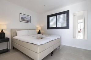 Une chambre à coucher avec un lit, deux tables de chevet avec des lampes, une fenêtre avec des stores, un tableau encadré au mur et un miroir sur le mur.