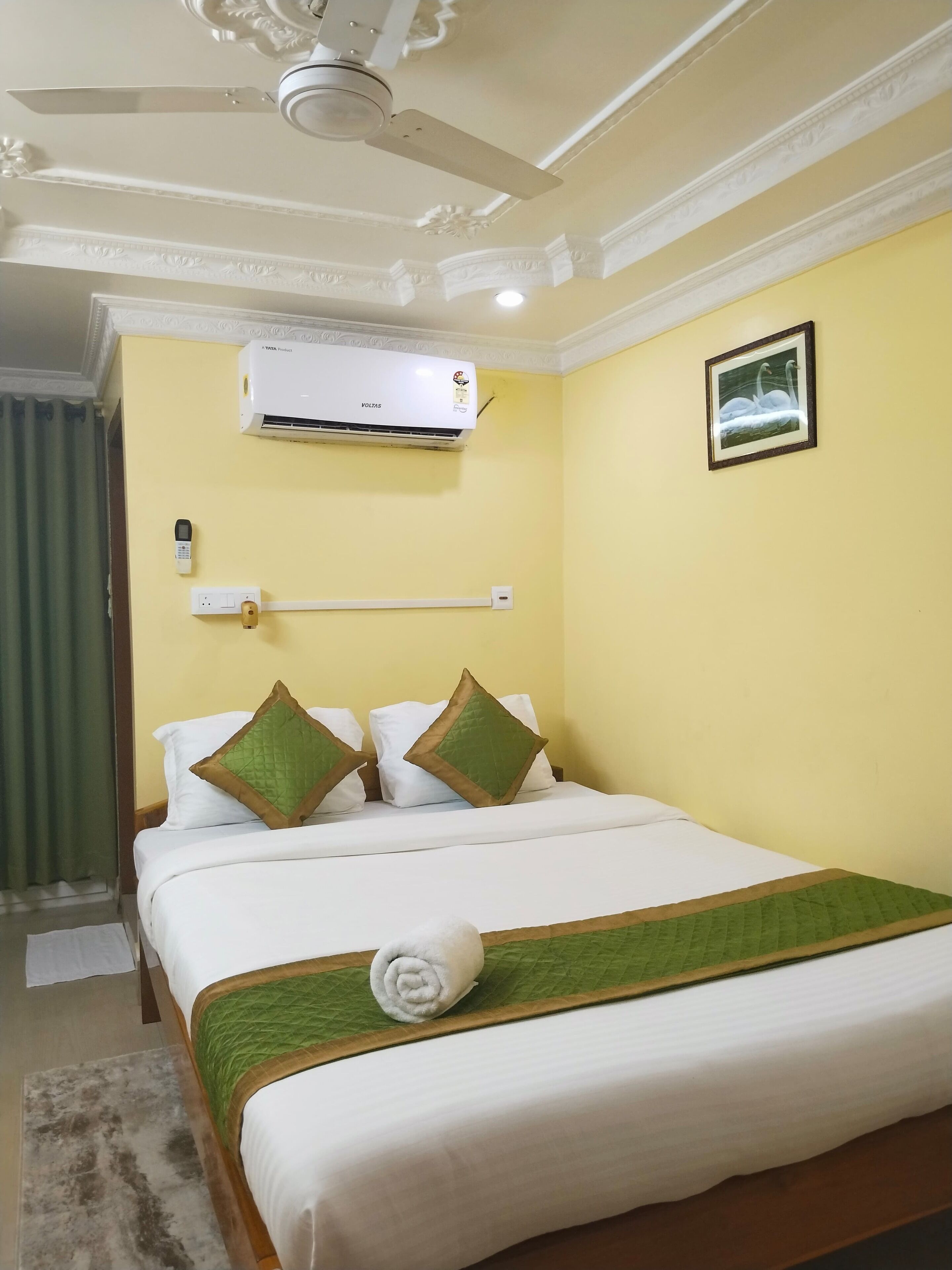 Habitación de hotel con cama, ventilador de techo, aire acondicionado, interruptor de luz en la pared y un cuadro con cisnes.