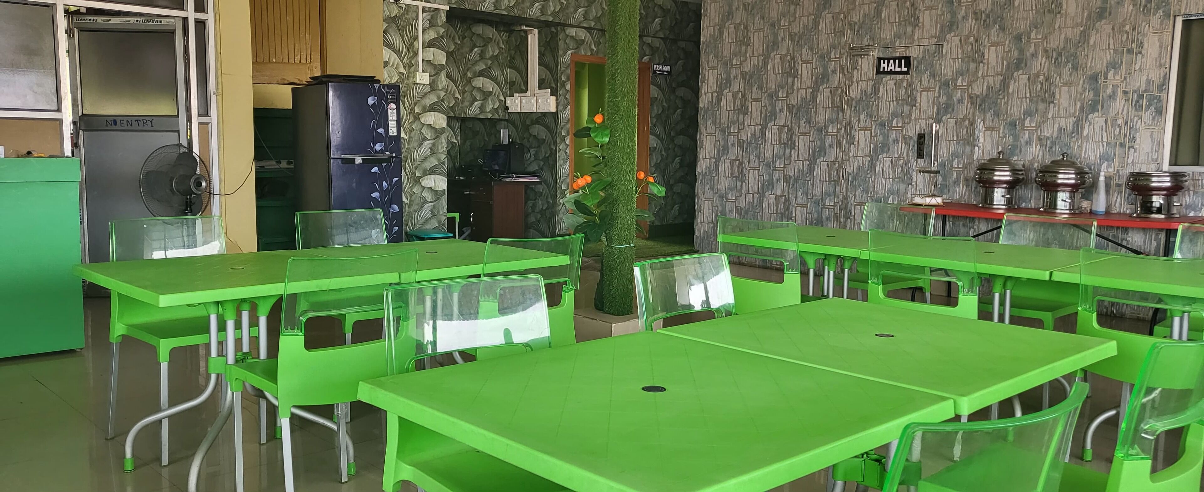 A dining area with green tables and chairs, a refrigerator, a fan, and a wall with a 'HALL' sign.