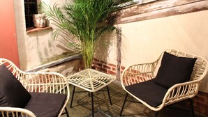 Een klein terras met rieten stoelen en een tafel, omgeven door planten en een bakstenen muur.