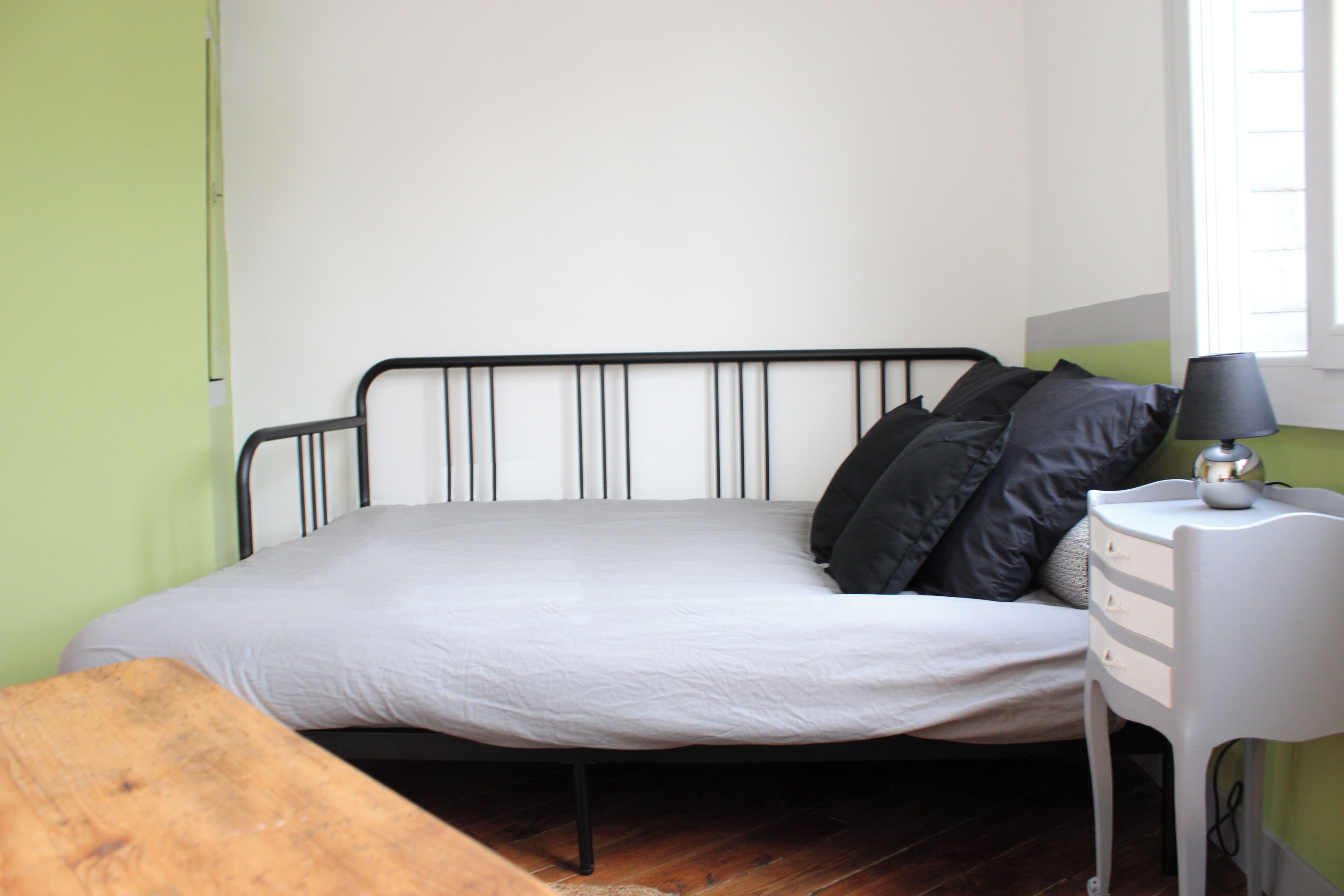 Ein Bett mit weißer Matratze und schwarzem Kopfteil, ein Nachttisch mit Lampe und eine Holzbank.