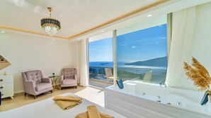 Una habitación de hotel moderna con una cama grande, dos sillones, un balcón con cerramiento de vidrio y vistas al mar, y una araña.