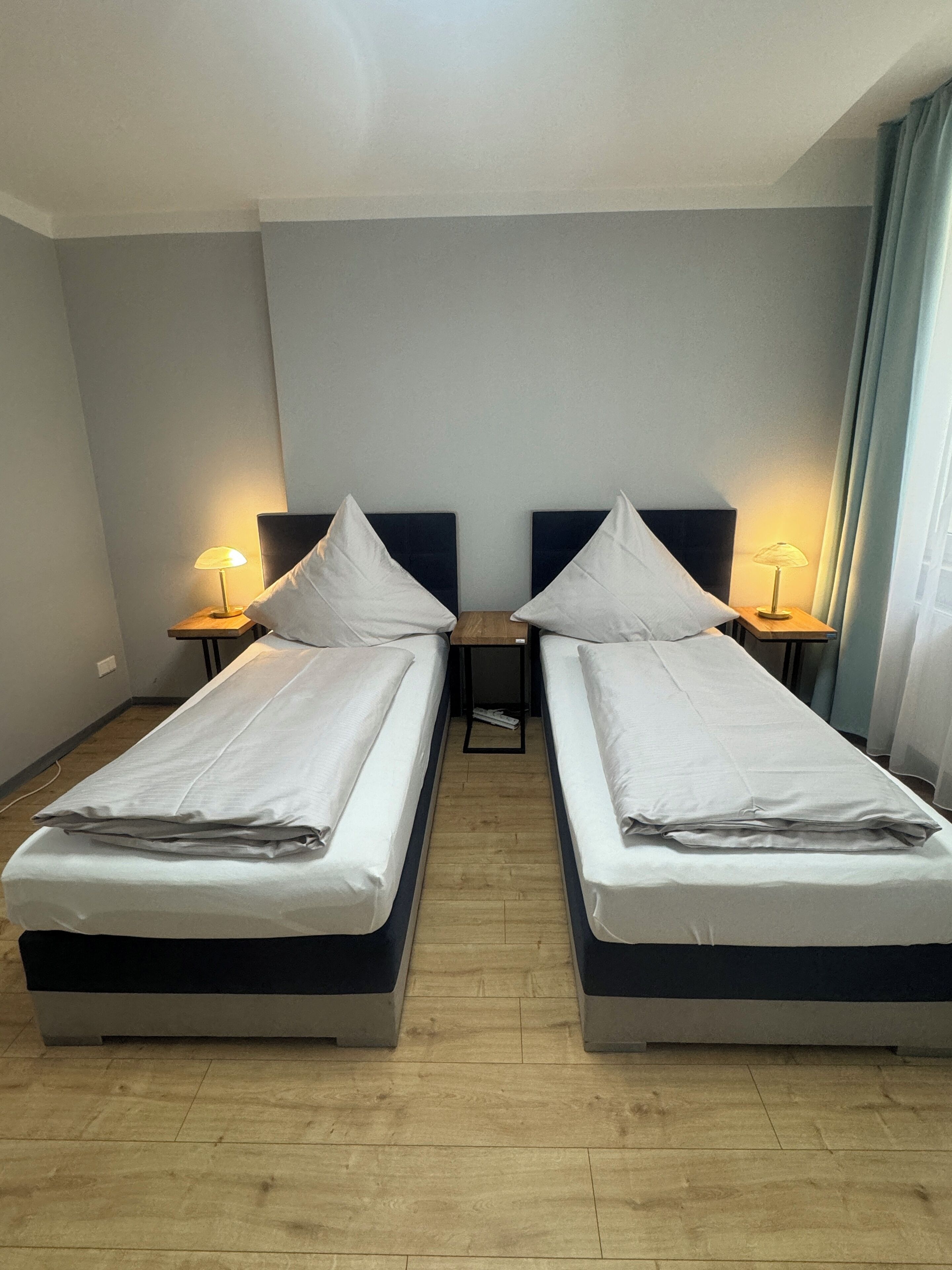 Ein Hotelzimmer mit zwei Einzelbetten, jeweils mit Holz Kopfteil und Nachttisch mit Lampe. Der Raum hat Holzboden und helle Wände.