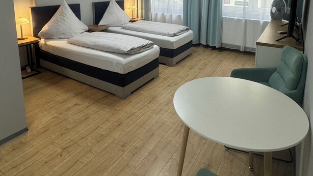 Quarto de hotel com duas camas, uma mesa redonda, uma cadeira, uma TV e uma janela com cortinas.