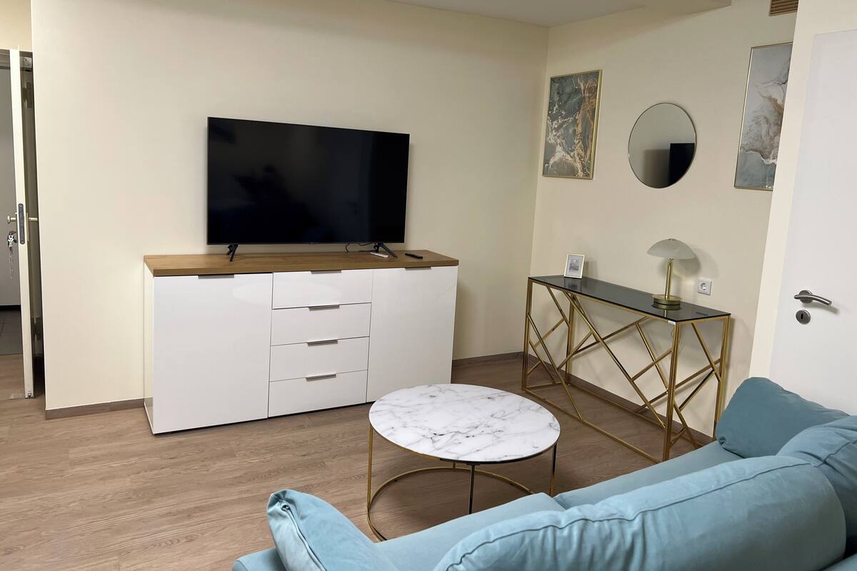 Un salon moderne avec un téléviseur à écran plat, un meuble blanc, une table ronde en marbre, un canapé bleu et une petite table d’appoint avec une lampe.