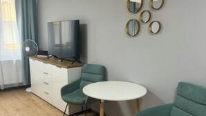 Uma sala de estar moderna com televisão de tela plana, uma mesa redonda branca, poltronas verde-água, um armário branco e um espelho fixado na parede.