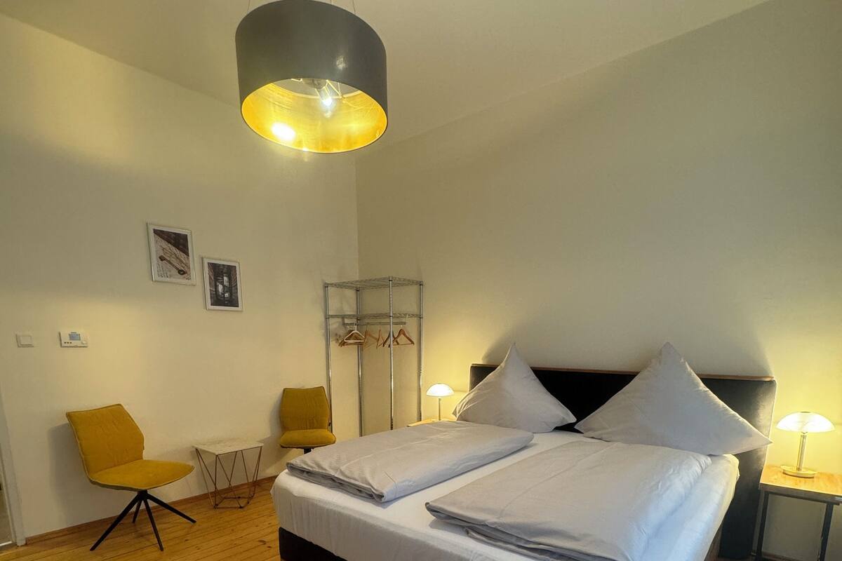 Une chambre avec un plancher en bois, un lit recouvert de draps blancs, deux chaises, une table de chevet, une lampe et un luminaire suspendu.