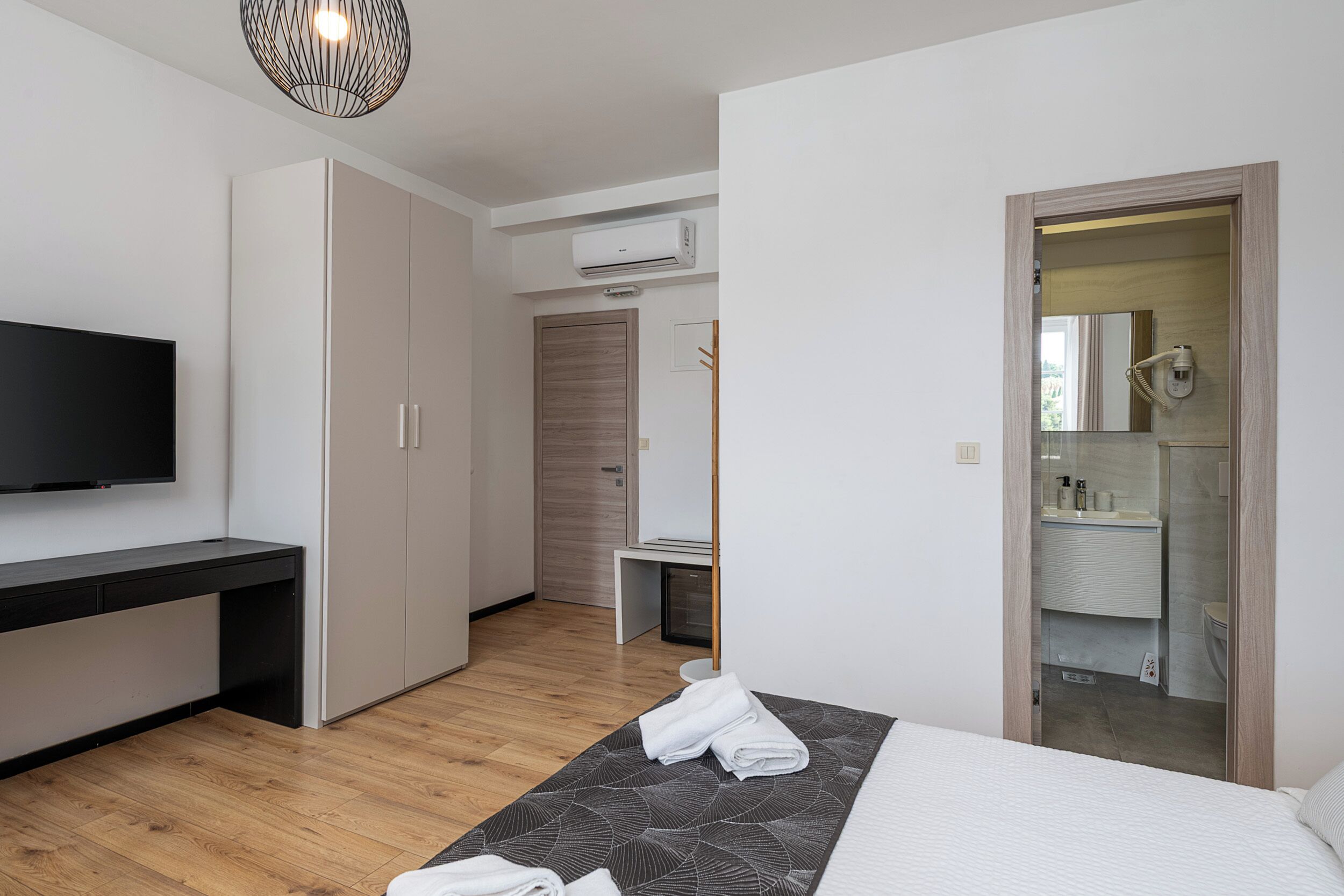 Una habitación de hotel moderna con televisión de pantalla plana, una cama con cubrecama estampado geométrico, suelo de madera, un armario y un baño con espejo y lavamanos.