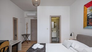 Quarto de hotel moderno com cama, escrivaninha e cadeira. Há um quadro na parede, uma porta que dá para o banheiro e um aparelho de ar condicionado.