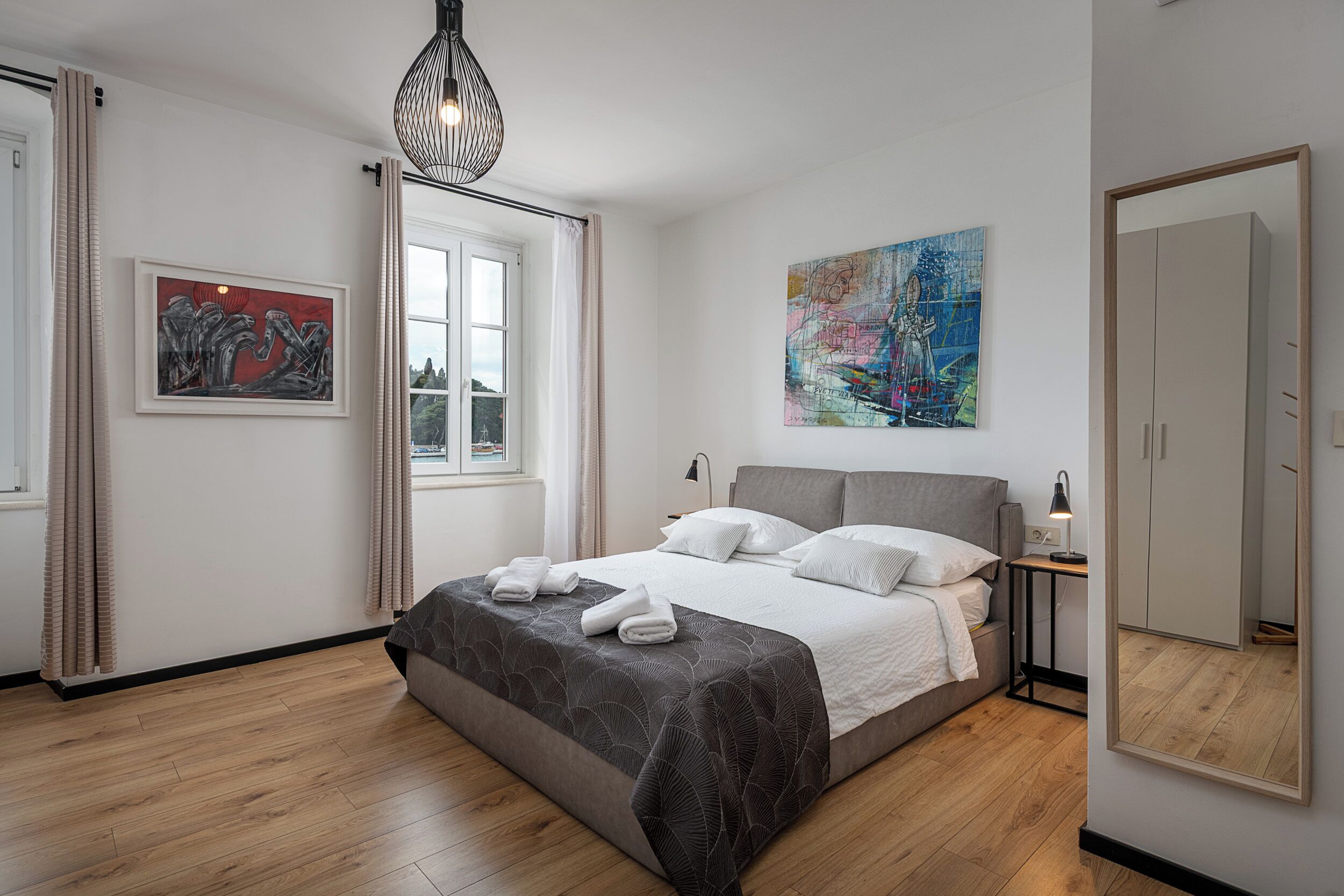 A bedroom with a bed, a large mirror, a map artwork, and a window with curtains.