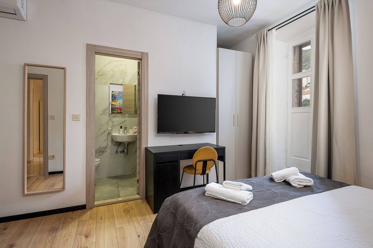 Una habitación de hotel moderna con cama, escritorio, silla, televisor, baño y armario.