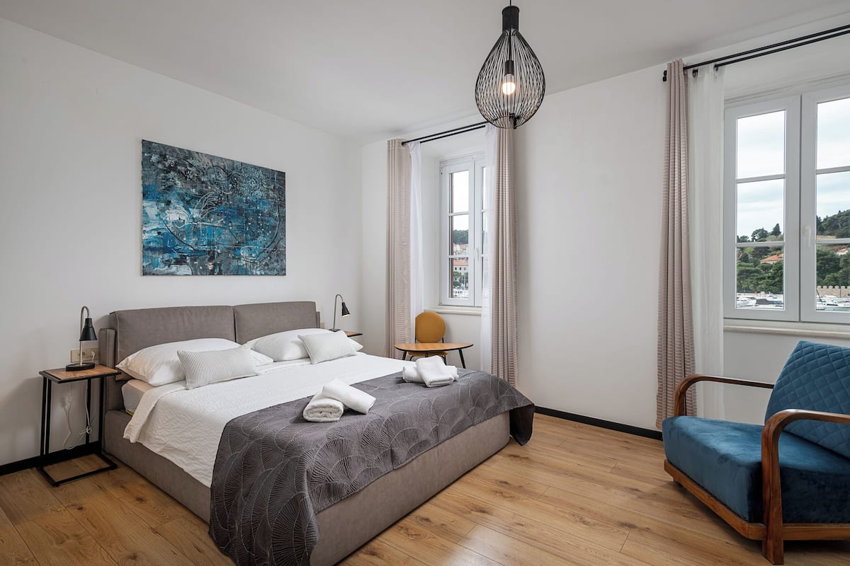 Un dormitorio moderno con una cama grande, una mesita de noche, una silla, un cuadro y un ventanal con cortinas.