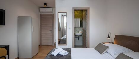 A modern hotel room with a bed, a desk, a wardrobe, and a bathroom visible through an open door.