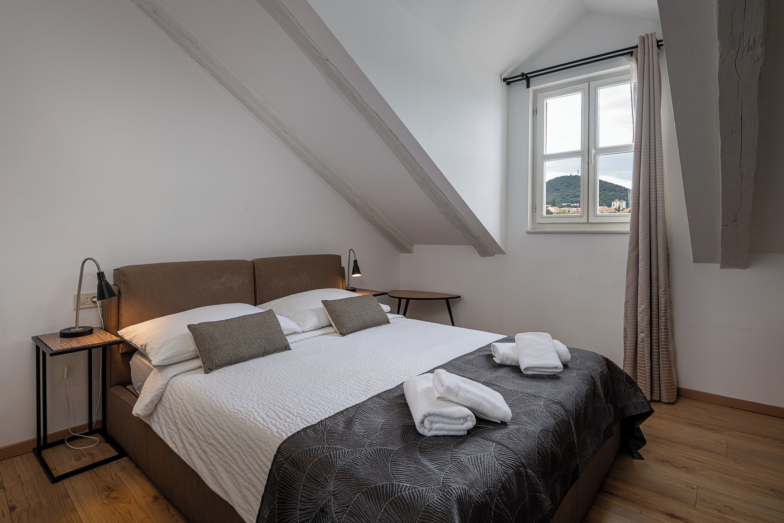A bedroom with a bed, bedside tables, a window with curtains, and a view of a hill.